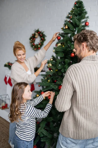 Chica sonriente decorando el árbol de Navidad cerca de papá y mamá borrosa en casa - foto de stock