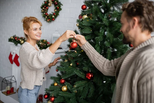 Mujer sonriente sosteniendo bauble cerca de marido y árbol de Navidad en casa - foto de stock