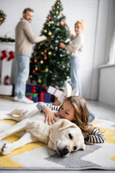 Smiling girl hugging labrador near burred parents and christmas tree at home — Stock Photo