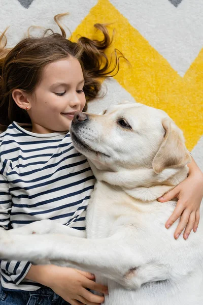 Visão superior do labrador abraçando menina sorridente no chão em casa — Fotografia de Stock