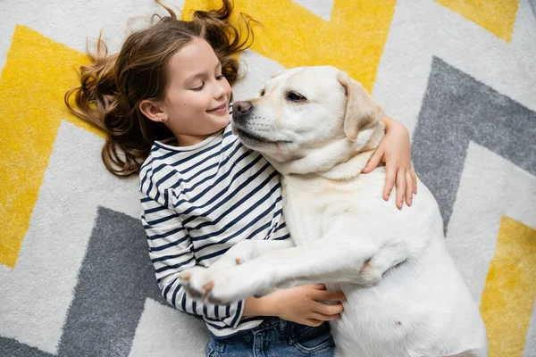 Draufsicht auf fröhliches Kind, das Labrador zu Hause auf dem Boden umarmt — Stockfoto