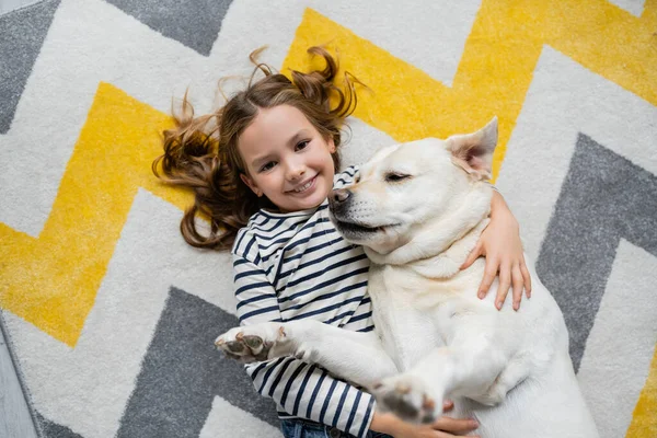 Draufsicht eines lächelnden Mädchens, das in die Kamera schaut und zu Hause Labrador auf dem Boden umarmt — Stockfoto