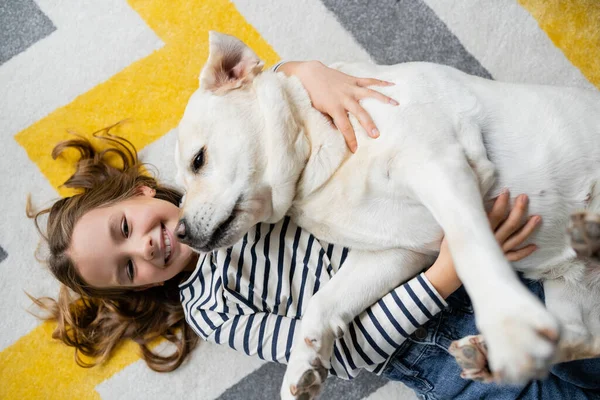 Visão superior da menina alegre abraçando labrador e olhando para a câmera no chão em casa — Fotografia de Stock