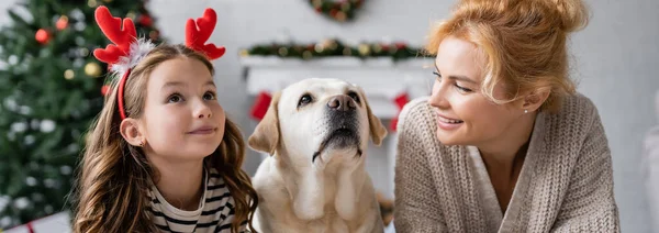 Positive woman looking at child with christmas headband near labrador at home, banner — Stock Photo