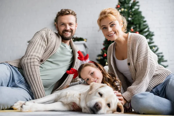 Positive parents looking at camera near daughter with christmas headband and labrador on floor at home — Stock Photo