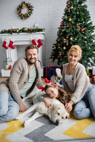 Familia positiva con hija y labrador sentados cerca del árbol de Navidad en casa - foto de stock