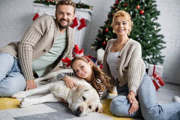 Sonriente familia y chica con diadema navideña mirando a la cámara cerca de labrador en el suelo en la sala de estar - foto de stock