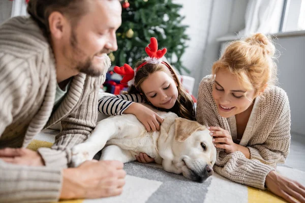 Positive mother and daughter petting labrador near blurred father at home during christmas — Stock Photo
