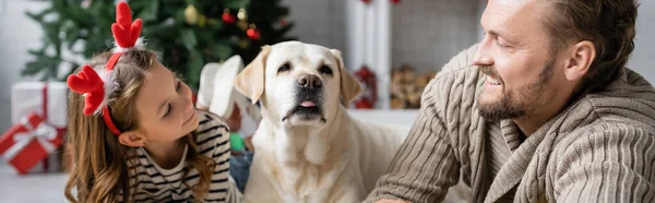 Homme joyeux regardant fille avec bandeau de Noël près de labrador à la maison, bannière — Photo de stock
