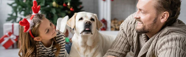 Hombre alegre mirando a la hija con diadema de Navidad cerca de labrador en casa, pancarta — Stock Photo