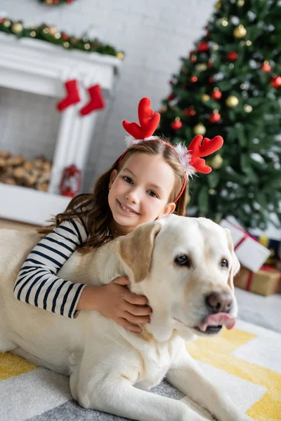 Sorrindo menina no Natal headband petting labrador em casa — Fotografia de Stock