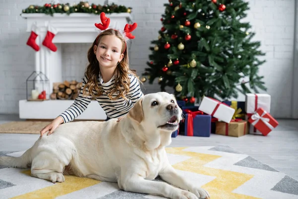 Mädchen mit Weihnachts-Stirnband blickt in die Kamera bei Labrador im Wohnzimmer — Stockfoto