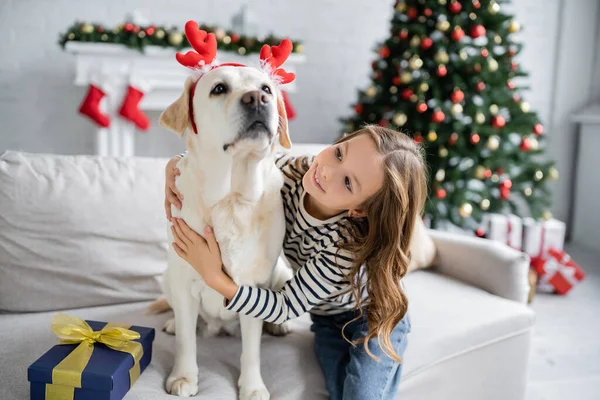 Kid abraçando labrador com headband de Natal perto presente no sofá em casa — Stock Photo