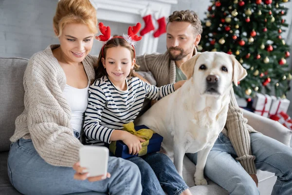 Donna positiva utilizzando il cellulare vicino figlia in possesso di regalo di Natale e marito con labrador a casa — Foto stock