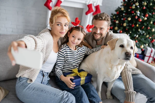 Familia sonriente con regalo de Navidad y labrador tomando selfie en smartphone en casa - foto de stock