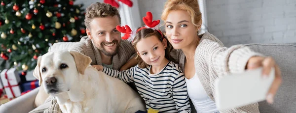 Famille positive avec labrador prendre selfie sur smartphone au cours de la nouvelle année à la maison, bannière — Photo de stock