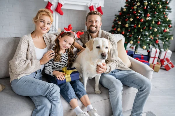 Smiling parents hugging daughter with christmas present and labrador at home — Stock Photo