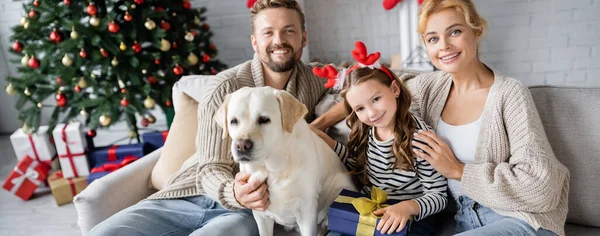 Fröhliche Familie mit Neujahrsgeschenk streichelt Labrador und schaut zu Hause in die Kamera, Banner — Stockfoto