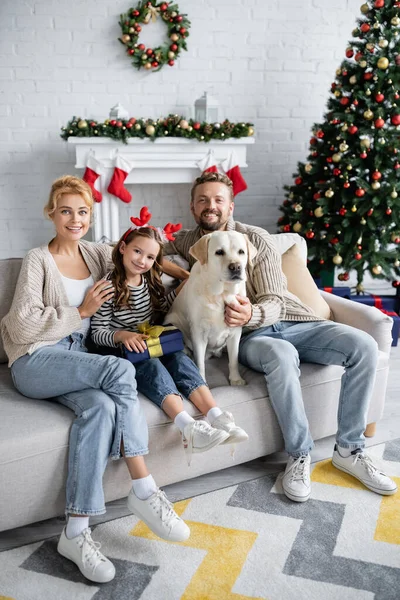 Pais abraçando a filha com presente e labrador durante a celebração do Natal em casa — Fotografia de Stock