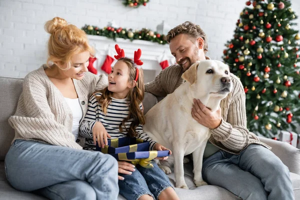 Chica abriendo regalo de Navidad cerca de los padres y labrador en casa - foto de stock