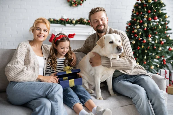 Girl opening christmas present near parents and labrador on couch — Stock Photo