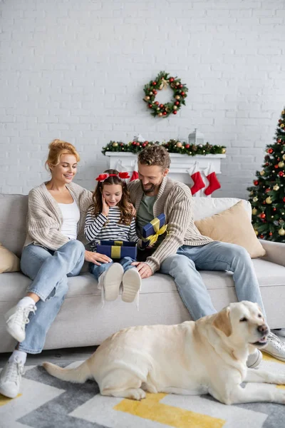 Enfant excité regardant ouvert boîte cadeau près des parents et labrador lors de la célébration de la nouvelle année à la maison — Photo de stock