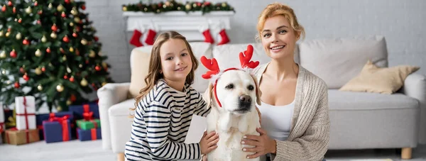 Alegre hija y mamá mirando a la cámara mientras acaricia labrador con diadema de Navidad en casa, pancarta — Stock Photo