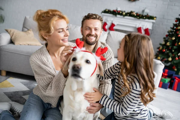 Positive Familie streichelt verschwommenen Labrador mit Weihnachts-Stirnband im Wohnzimmer — Stockfoto