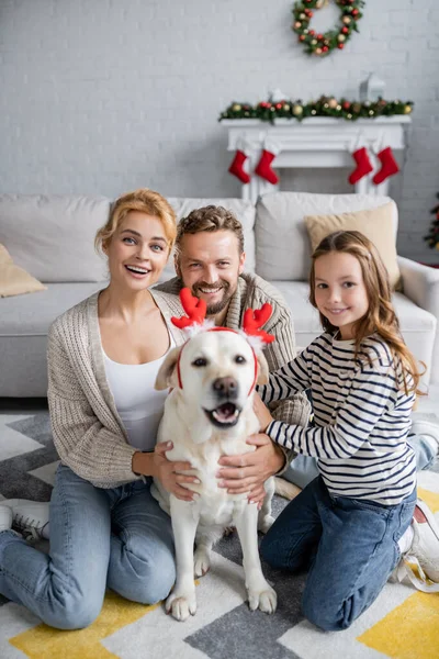 Familia feliz mirando a la cámara cerca borrosa labrador con diadema de Navidad en la sala de estar - foto de stock