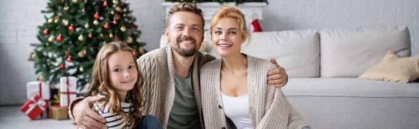 Sonriente hombre abrazando esposa e hija durante la Navidad en casa, pancarta - foto de stock