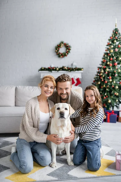 Pais felizes e menina olhando para a câmera perto labrador no tapete e árvore de natal borrada em casa — Fotografia de Stock