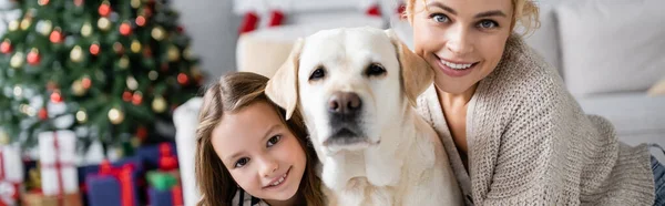 Donna e figlia guardando la fotocamera vicino labrador durante il nuovo anno a casa, banner — Foto stock