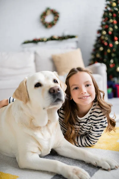 Lächelndes Mädchen umarmt Labrador, während es zu Neujahr auf dem Boden liegt — Stockfoto