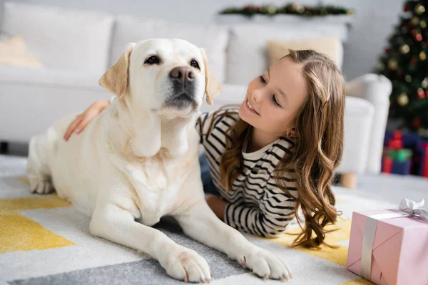 Happy kid hugging labrador near new year present on floor at home — Stock Photo