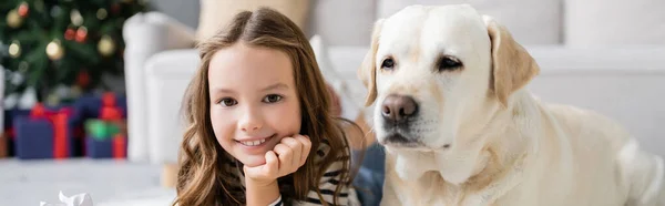 Niño sonriendo a la cámara cerca de labrador en casa, pancarta - foto de stock