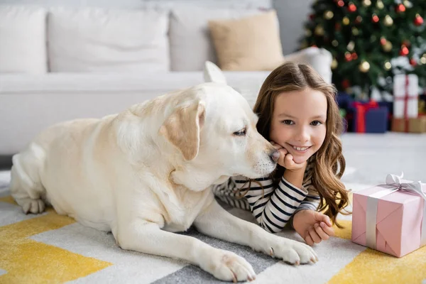 Labrador couché près d'enfant souriant et cadeau de Noël sur le tapis à la maison — Photo de stock