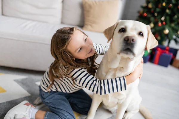 Chica sonriente abrazando labrador borrosa en casa durante el año nuevo - foto de stock