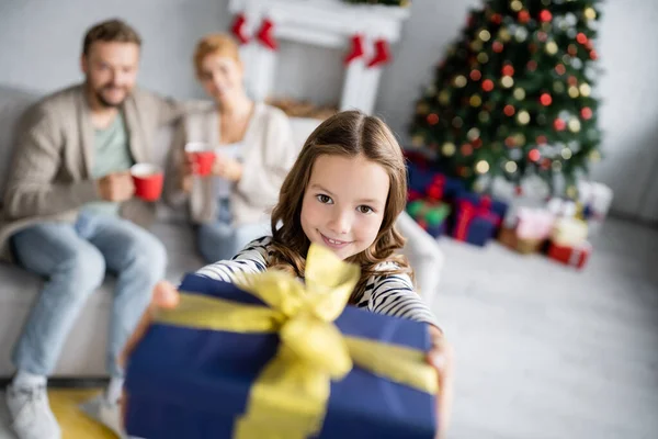 Fröhliches Mädchen hält Weihnachtsgeschenk in der Hand und blickt in die Kamera neben verschwommenen Eltern im Wohnzimmer — Stockfoto