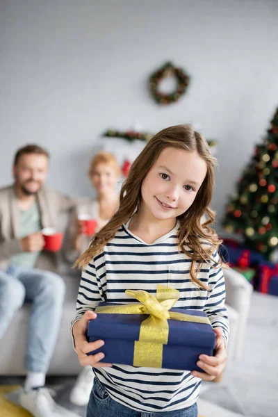 Kid holding new year gift near blurred family at home — Stock Photo