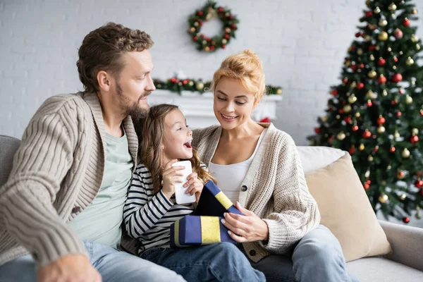 Smiling woman opening gift box with smartphone during christmas at home — Stock Photo