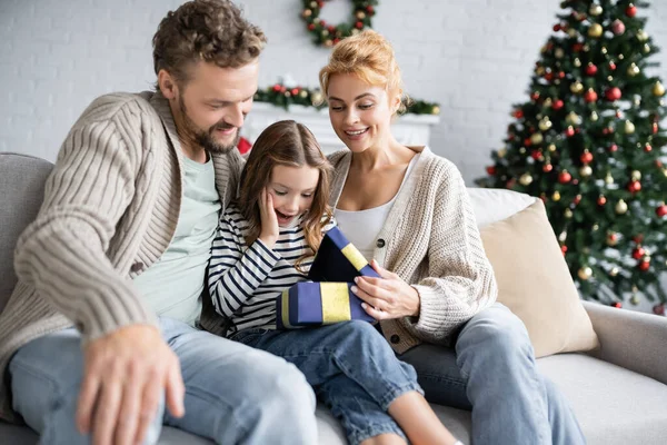 Chica emocionada mirando la caja de regalo abierta cerca de los padres durante la Navidad en casa - foto de stock