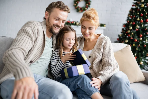 Sonrientes padres mirando a la hija abriendo regalo de Navidad en el sofá en casa - foto de stock