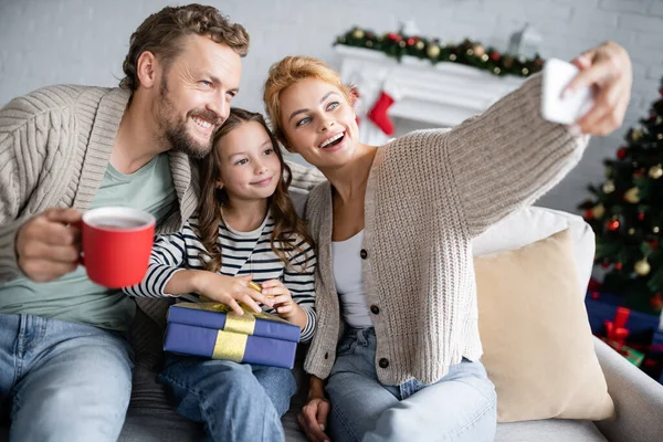 Woman taking selfie with family holding christmas gift and cup on couch at home — Stock Photo