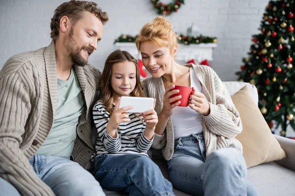 Souriant fille en utilisant smartphone près de la mère avec tasse et papa sur le canapé pendant la nouvelle année — Photo de stock