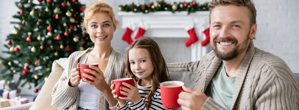 Joyeux famille tenant des tasses de cacao sur le canapé pendant Noël à la maison, bannière — Photo de stock