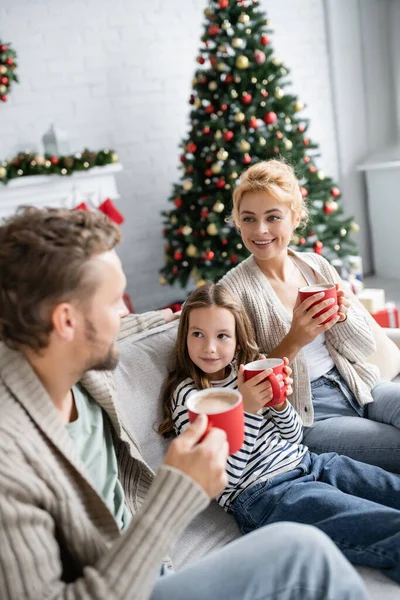 Sonriente madre y niña mirando al borroso padre con taza de cacao durante la Navidad en casa - foto de stock