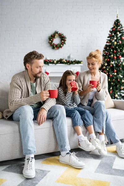 Menina bebendo cacau perto dos pais no sofá durante o Natal em casa — Fotografia de Stock