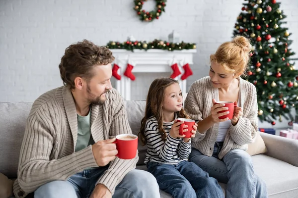 Lächelnde Eltern schauen Tochter beim Neujahrsempfang mit Kakao auf Couch an — Stockfoto