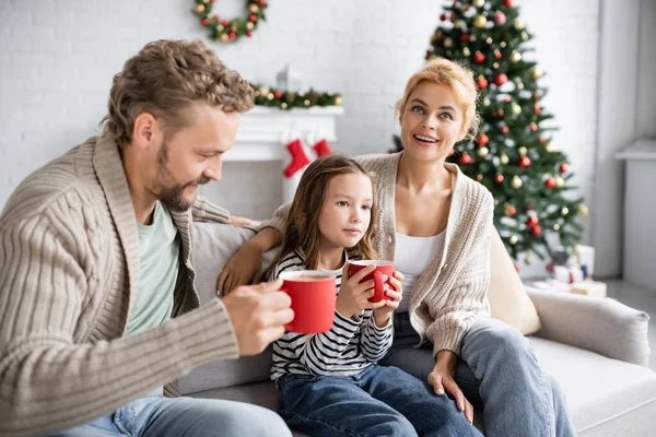 Mujer sonriente hablando con la familia con tazas de cacao en el sofá durante la Navidad - foto de stock