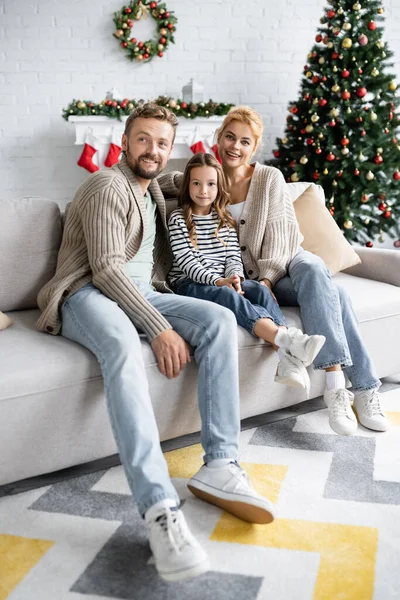 Lächelnde Familie blickt in die Kamera, während sie auf der Couch in der Nähe des verschwommenen Weihnachtsbaums sitzt — Stockfoto